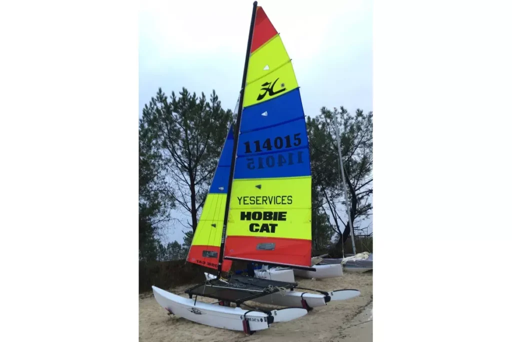 Catamaran Hobie Cat 16 blanc à voile colorée (jaune, bleu, rouge) marqué YESERVICES 114015 sur une plage de sable.
