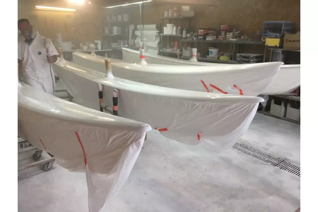 Technicien en combinaison pulvérisant de la peinture sur des coques de bateaux protégées dans un atelier nautique.