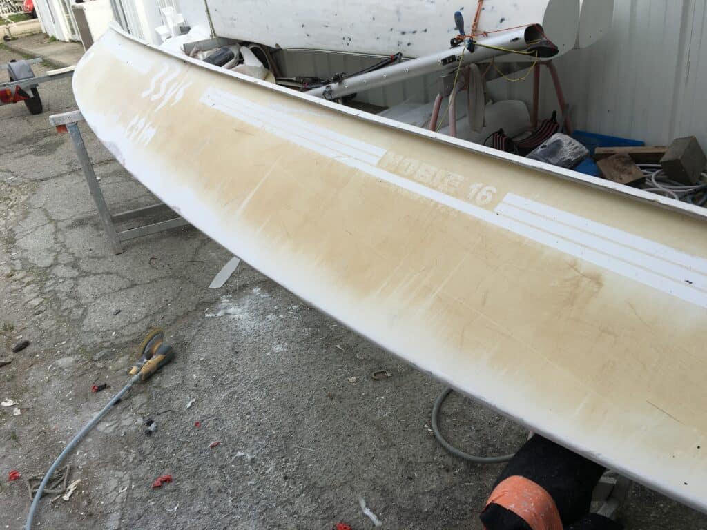 Restauration et ponçage d'une coque de voilier Hobie 16 Restauration d'une coque de Hobie 16 jaunie avec ponceuse électrique au sol. Texte visible : HOBIE 16 et 33116.