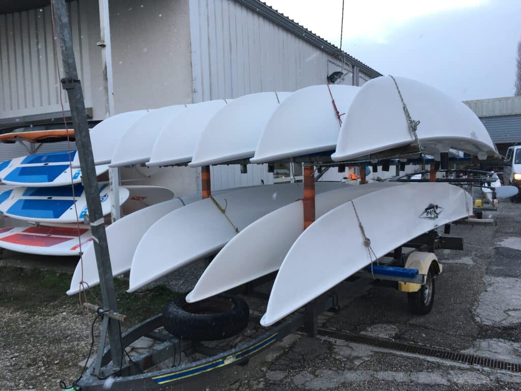 Stockage et Transport de Catamarans Hobie sur Remorque Catamarans Hobie blancs empilés sur une remorque de transport devant un hangar sous un ciel gris.