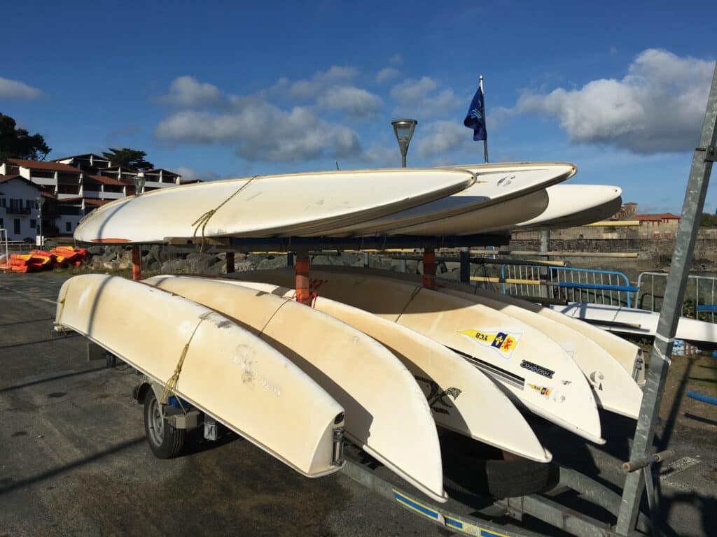 Voiliers dériveurs blancs sur remorque au bord de mer Dériveurs blancs empilés sur une remorque au bord de l'eau avec logos Tribord et YCB sous un ciel bleu nuageux.