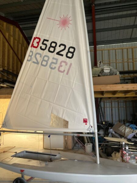 Voilier Laser blanc avec voile numérotée 135828 et logo rose, entreposé dans un hangar sur sa remorque.