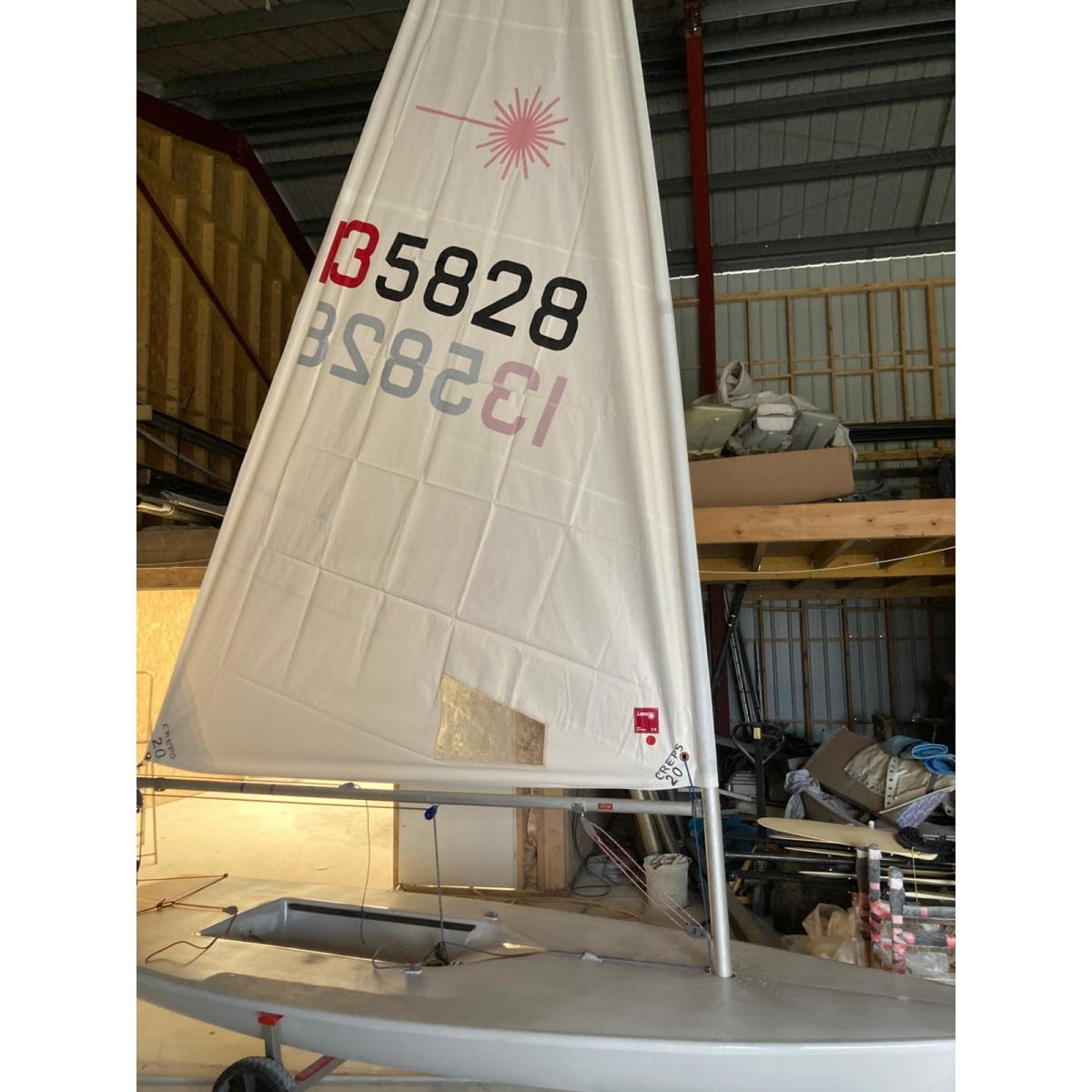 Voilier Laser blanc avec voile numérotée 135828 et logo rose, entreposé dans un hangar sur sa remorque.